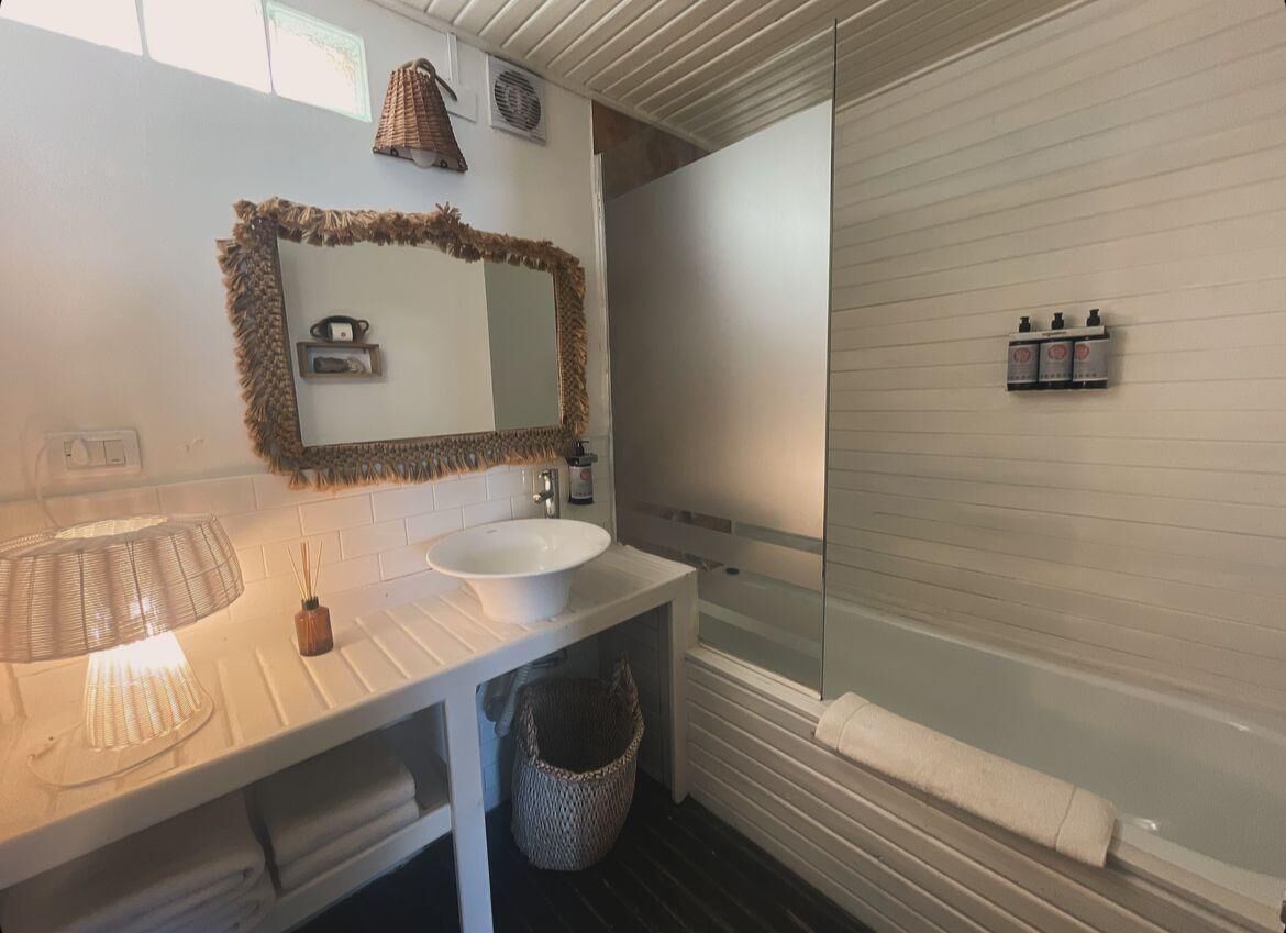 Un baño con lavamanos blanco, espejo y ducha. Hay artículos de aseo en la pared y una toalla en el suelo.