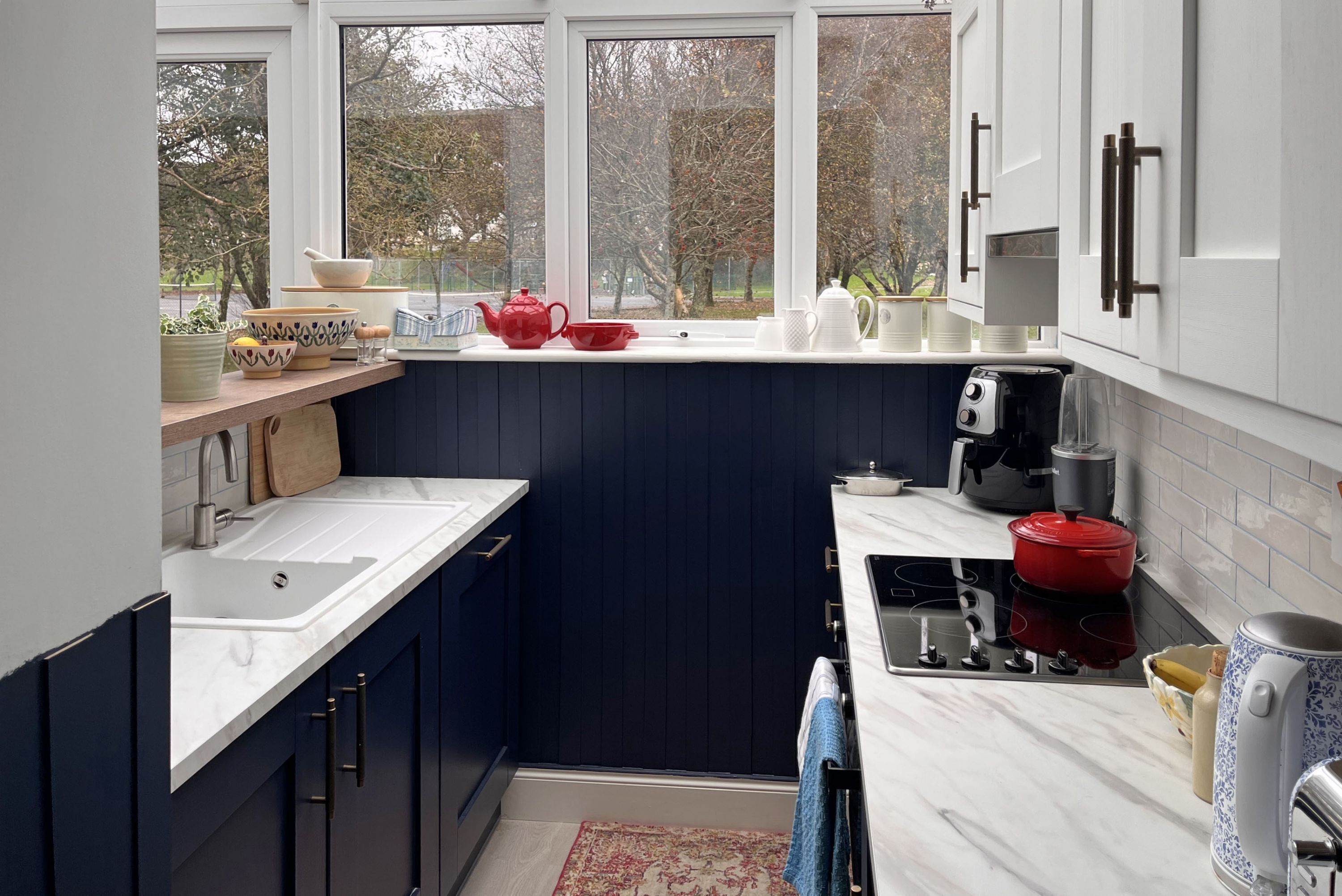 Una cocina moderna con armarios azul oscuro, encimeras blancas y ventana con vistas a un jardín.