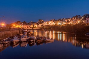 Un poble al costat del mar a la posta de sol, amb vaixells amarrats que reflecteixen els edificis i els fanals.