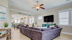 Un salon moderne avec un grand canapé d’angle, un ventilateur de plafond, un téléviseur fixé au mur et un coin cuisine à l’arrière-plan.