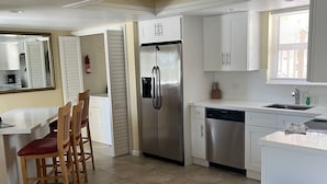 Cocina compacta con refrigerador de acero inoxidable, lavavajillas y pileta. Se observan sillas de madera y una mesita, además de un basurero.