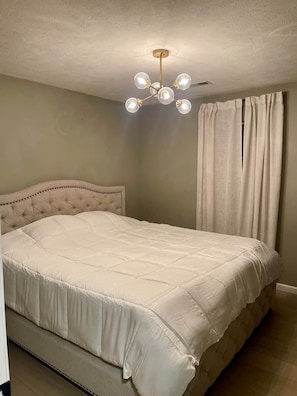 Une chambre à coucher avec une tête de lit capitonnée, un lit recouvert d’une courtepointe blanche matelassée, un luminaire au plafond et une fenêtre avec des rideaux.