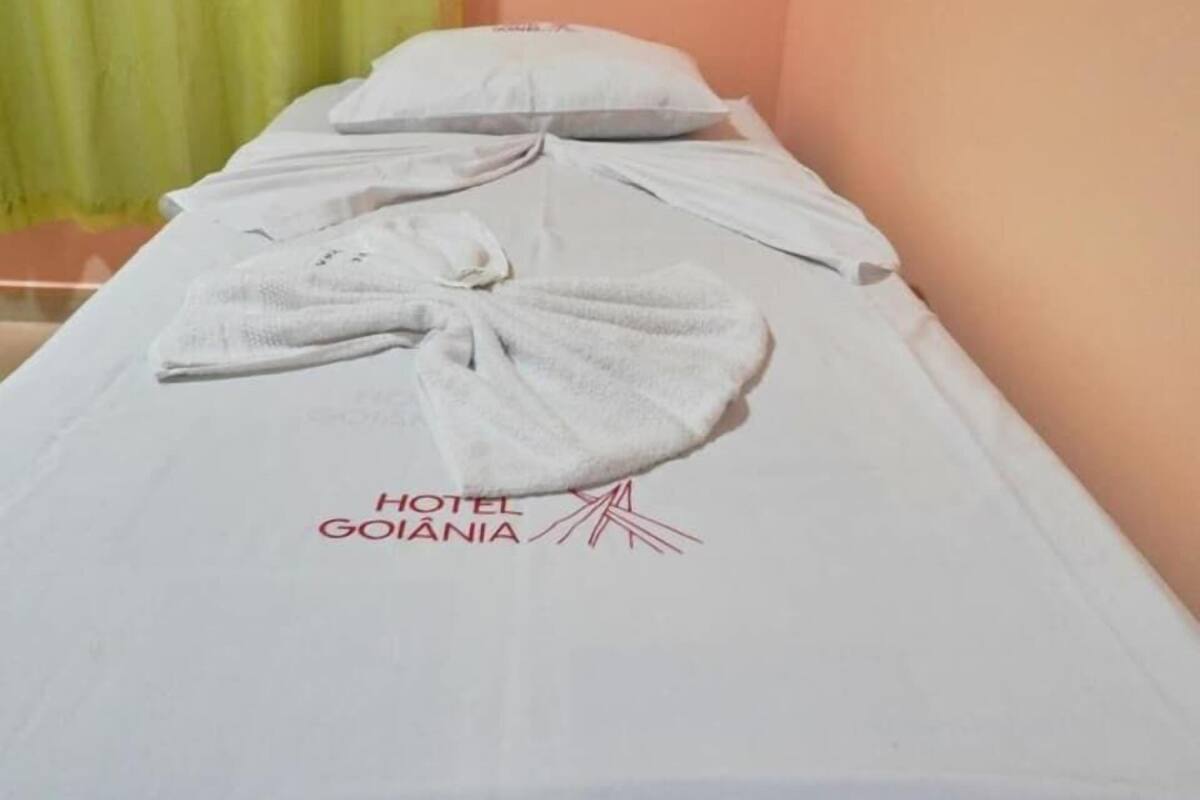 A single bed with a towel and pillow, a green curtain, and the text 'HOTEL GOIANIA' on the bedspread.