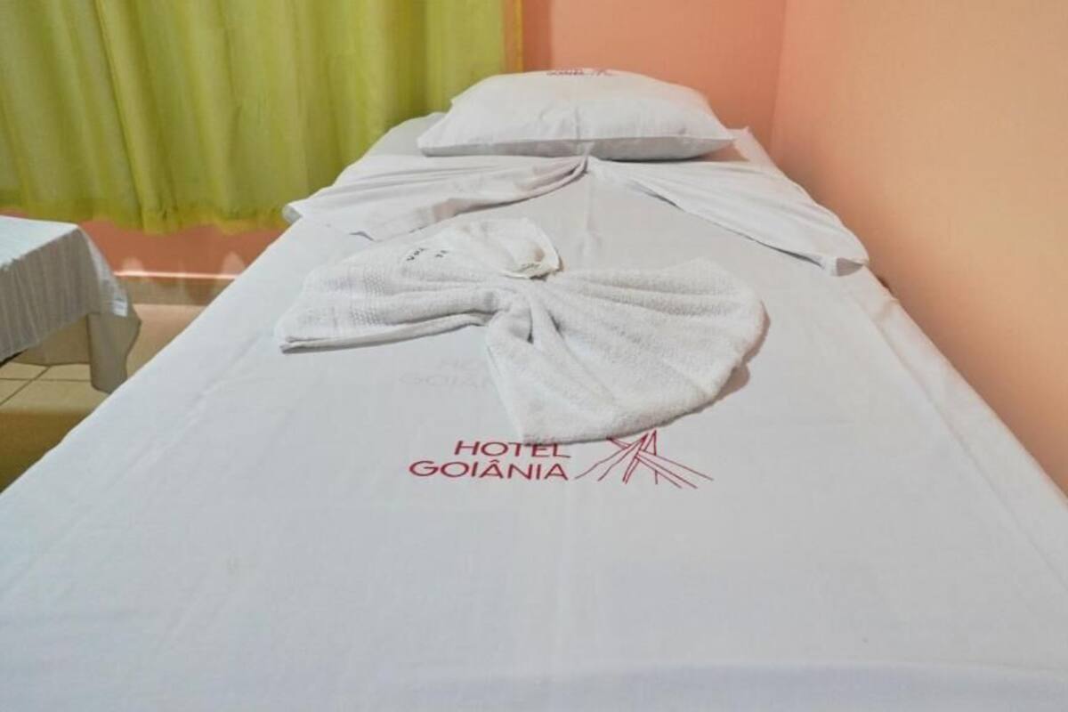 A hotel room with a neatly made bed, a pillow, and a towel arranged on the bed. The room has a simple layout with a plain wall and a curtain in the background.