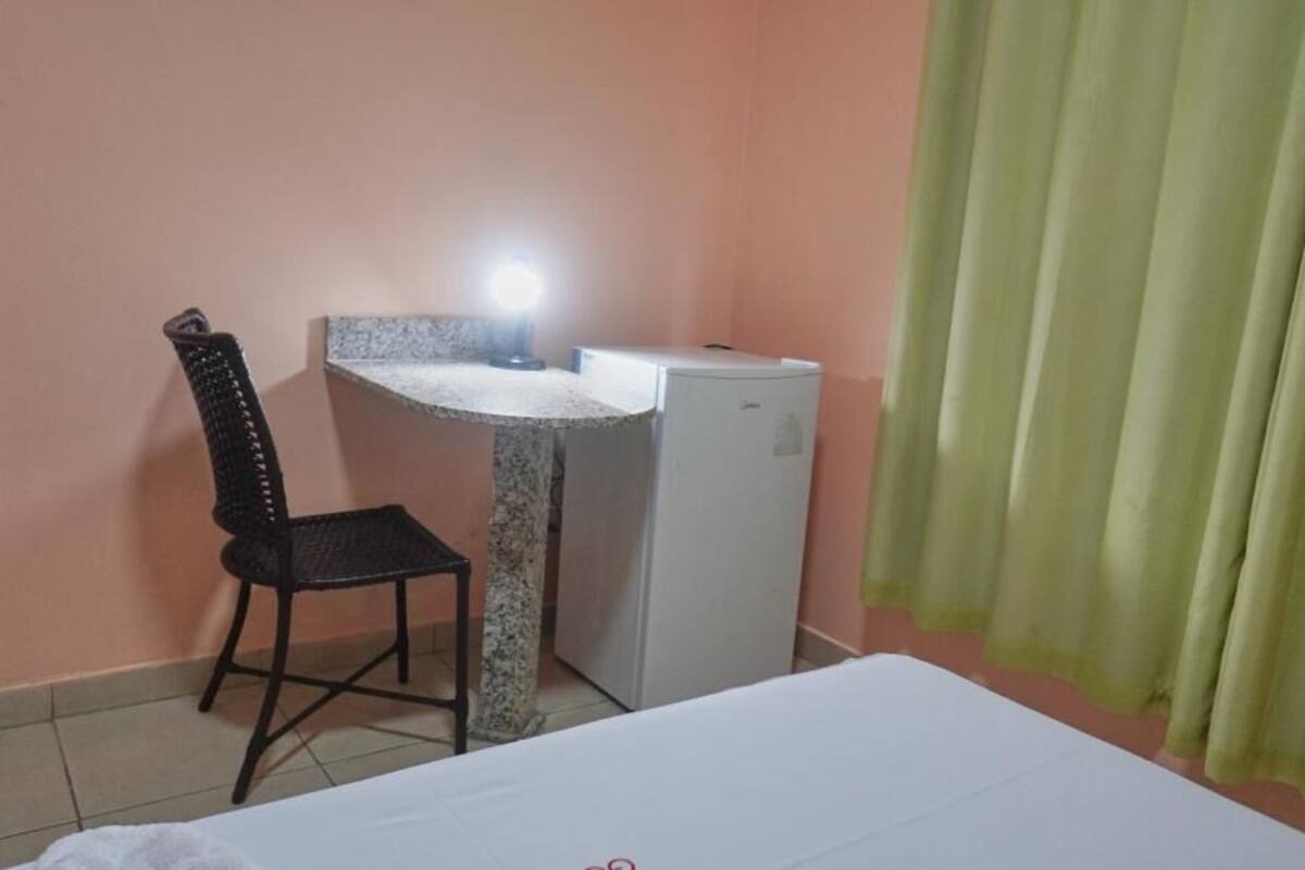 A small room with a bed, a desk with a chair, a refrigerator, and a window with curtains.