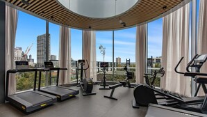 Une salle de sport moderne équipée de tapis de course, de vélos elliptiques et d’un rameur, et dotée de grandes fenêtres offrant une vue sur la ville.