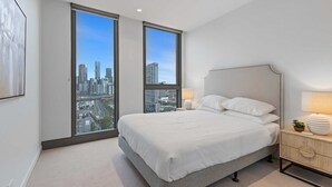 Une chambre moderne avec un grand lit, deux fenêtres donnant sur la ville, une table de chevet avec une lampe et une plante décorative.