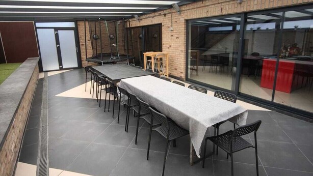 Uma área de refeições ao ar livre com uma mesa comprida e cadeiras, um banquinho de madeira pequeno e um muro de vidro com um objeto vermelho ao fundo.