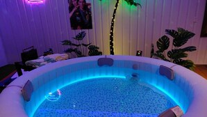 A hot tub with blue LED lighting, surrounded by potted plants, in a room with white walls and a poster.