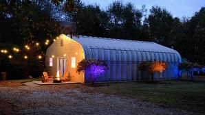A covered structure with a fire pit and seating area, surrounded by trees and string lights.