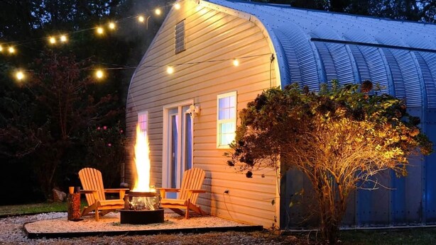 A small wooden house with a fire pit and two chairs outside, surrounded by a garden.