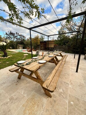 An outdoor dining area with a wooden picnic table set for four, surrounded by a pergola.