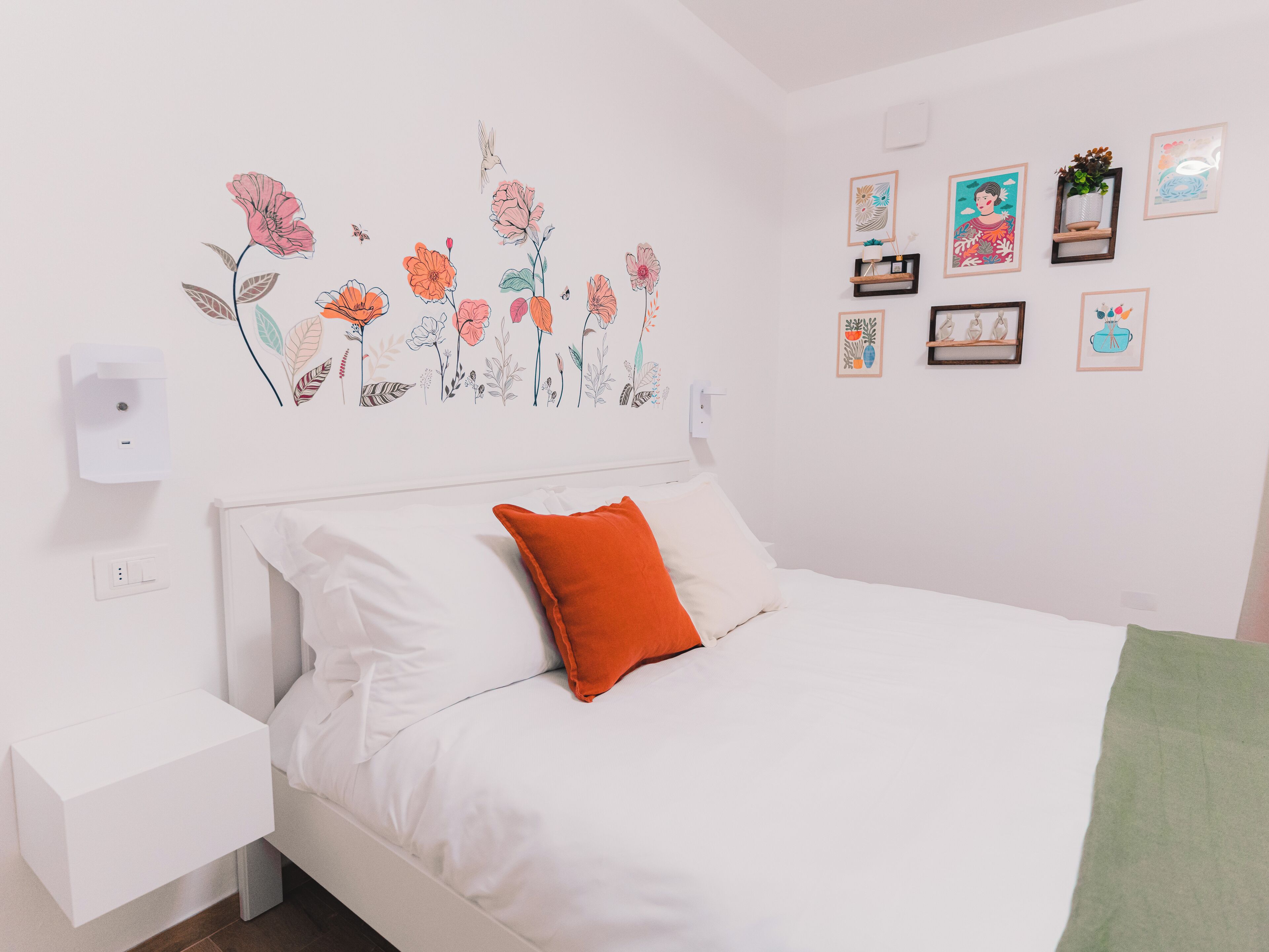 Un dormitorio con cama, ropa de cama blanca, un cojín naranja, una lámpara de pared y un adhesivo floral.