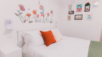 Ein Schlafzimmer mit einem Bett, weißer Bettwäsche, einem orangefarbenen Kissen, einer Wandleuchte und einer Blumensticker-Motiv.