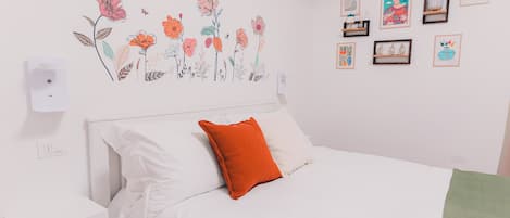 A bedroom with a bed, white bedding, an orange pillow, a wall-mounted light, and a floral decal.