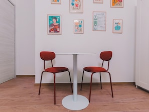 Une pièce avec une table blanche et deux chaises rouges, une porte blanche et un mur orné de tableaux encadrés.
