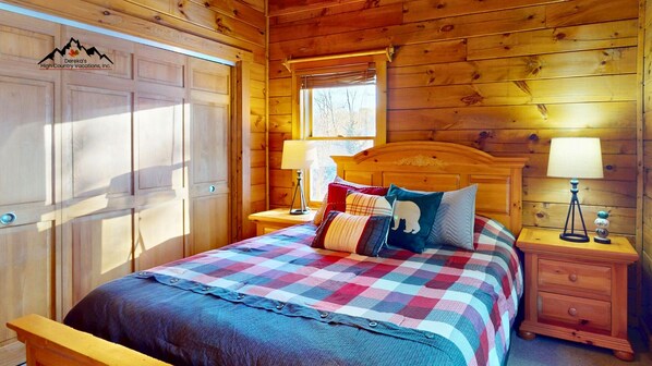 A wooden cabin bedroom with a bed, nightstands, a window, and a door. - Sweet Retreat • Hot Tub, Fire Pit, Pet Friendly (Sugar Mountain)