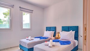 A hotel room with two beds, each with a blue headboard, white bedding, and towels. There are windows with blinds, a nightstand with a lamp, and a door. - Drastis Escape Villas - Villa Simran, By Estia (Peroulades)