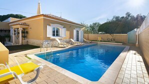 A swimming pool with lounge chairs, a tiled deck, and a building with a chimney. - Holiday Home with Shared Pool (Albufeira)