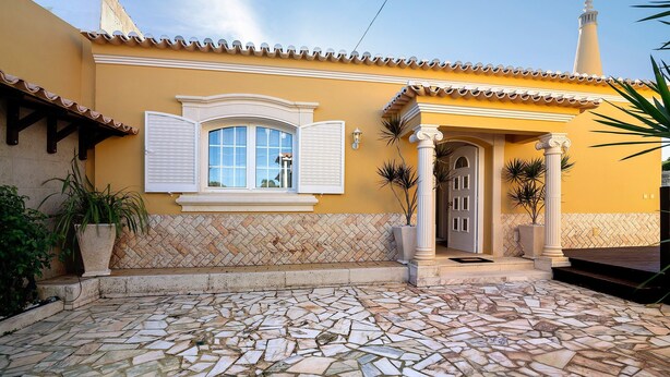 A yellow building with a tiled roof, a stone patio, and a window with white shutters. - Holiday Home with Shared Pool (Albufeira)