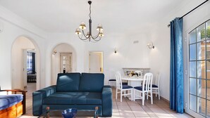 A spacious living room with a blue sofa, a glass coffee table, and a dining area with white chairs and a table. - Holiday Home with Shared Pool (Albufeira)