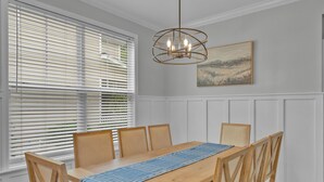 Un coin repas avec une table en bois, six chaises, un chemin de table bleu, des murs lambrés blancs, un luminaire au plafond et un tableau représentant un paysage.