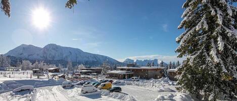 A snowy mountain landscape with a clear sky, a parking lot with several cars, and buildings with snow-covered roofs.