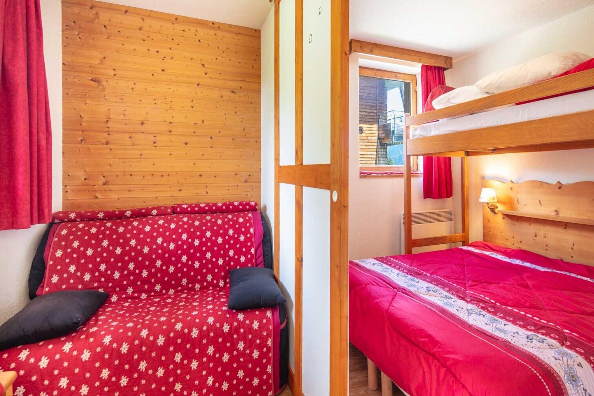 A room with a bunk bed, a sofa with a floral cover, and a window with red curtains.