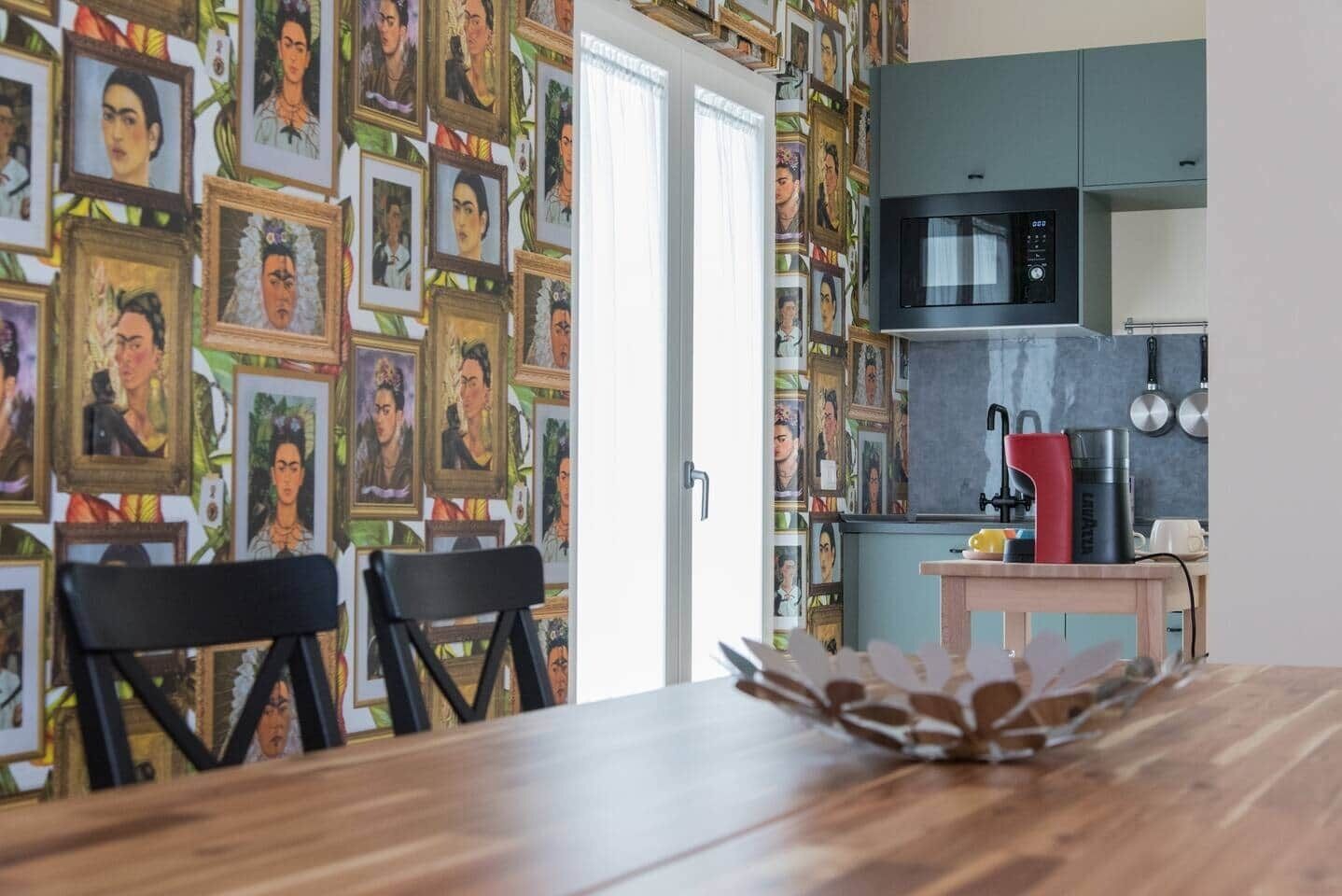 Un comedor con una mesa de madera, dos sillas y una pared decorada con retratos enmarcados. Se ve una cocina con microondas y hervidor en el fondo.
