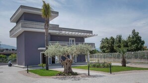 Un edificio moderno de dos pisos con fachada gris, un área de estar al aire libre cubierta y un jardín bien cuidado con árboles y arbustos.
