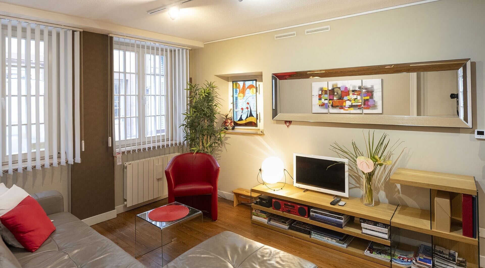 Un salon avec un canapé, un fauteuil rouge, un téléviseur, une bibliothèque et une fenêtre avec des stores.