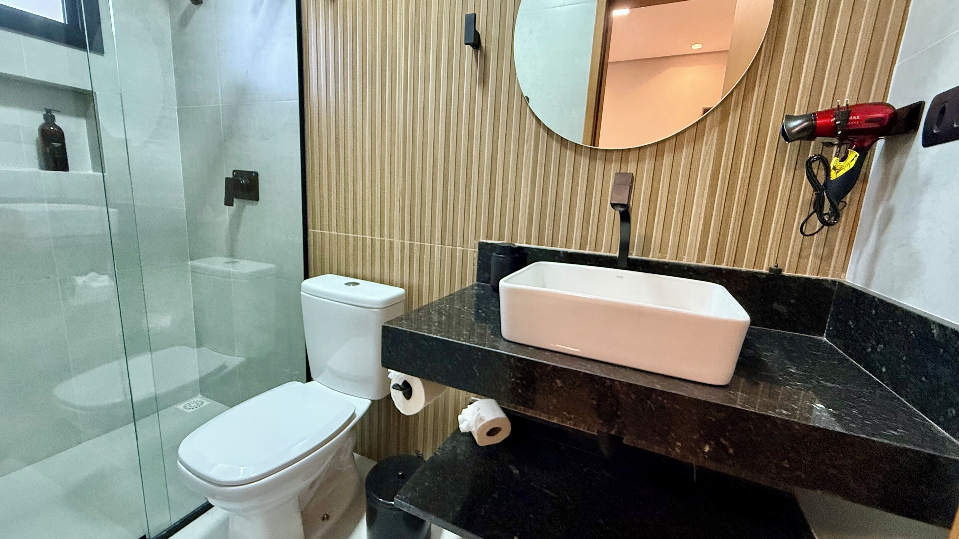 Um banheiro moderno com box de vidro, vaso sanitário branco, bancada preta com pia retangular branca e espelho sobre a pia.