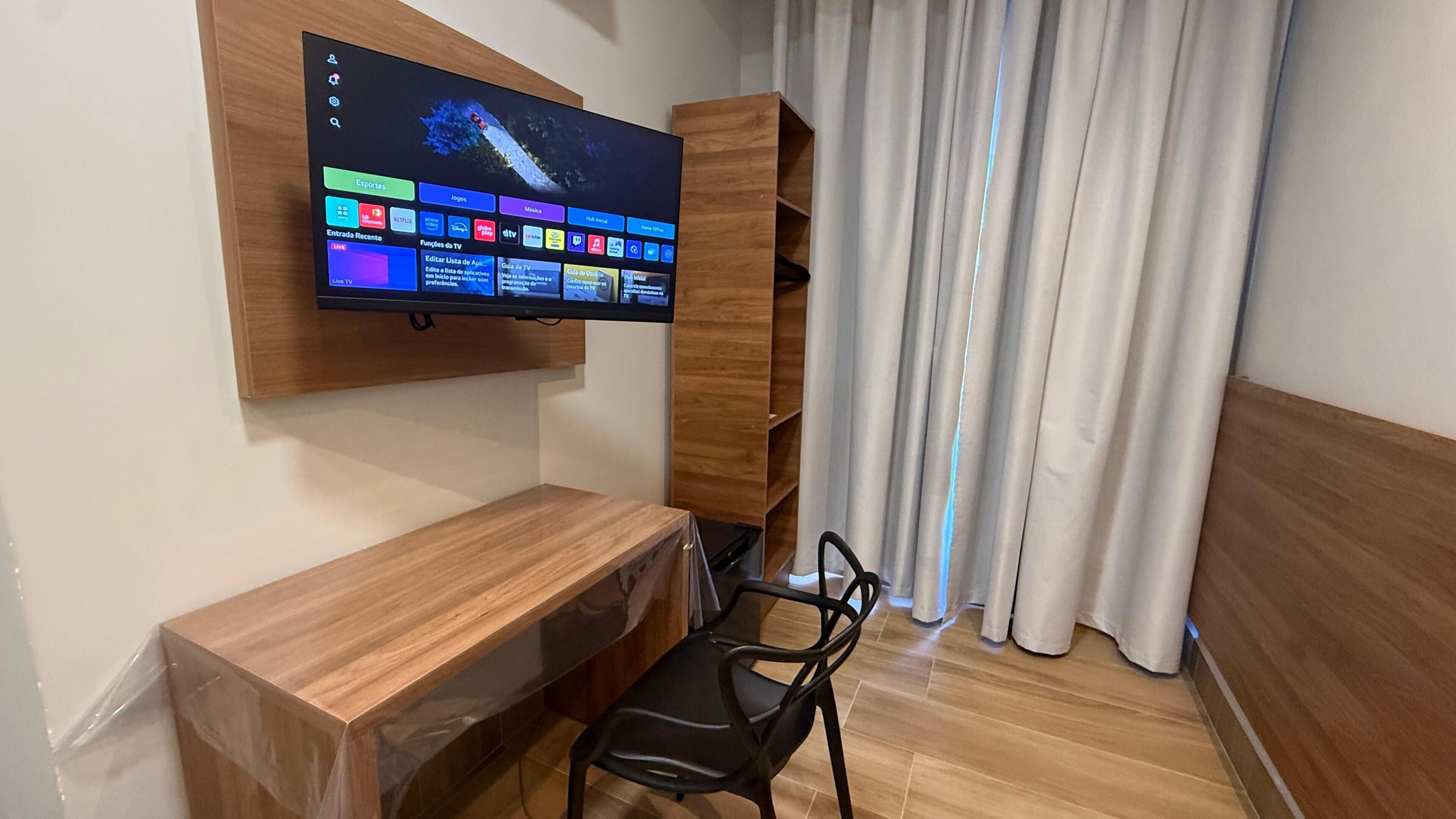 Quarto com escrivaninha de madeira, cadeira preta, TV fixada na parede exibindo guia de programação e cortina.