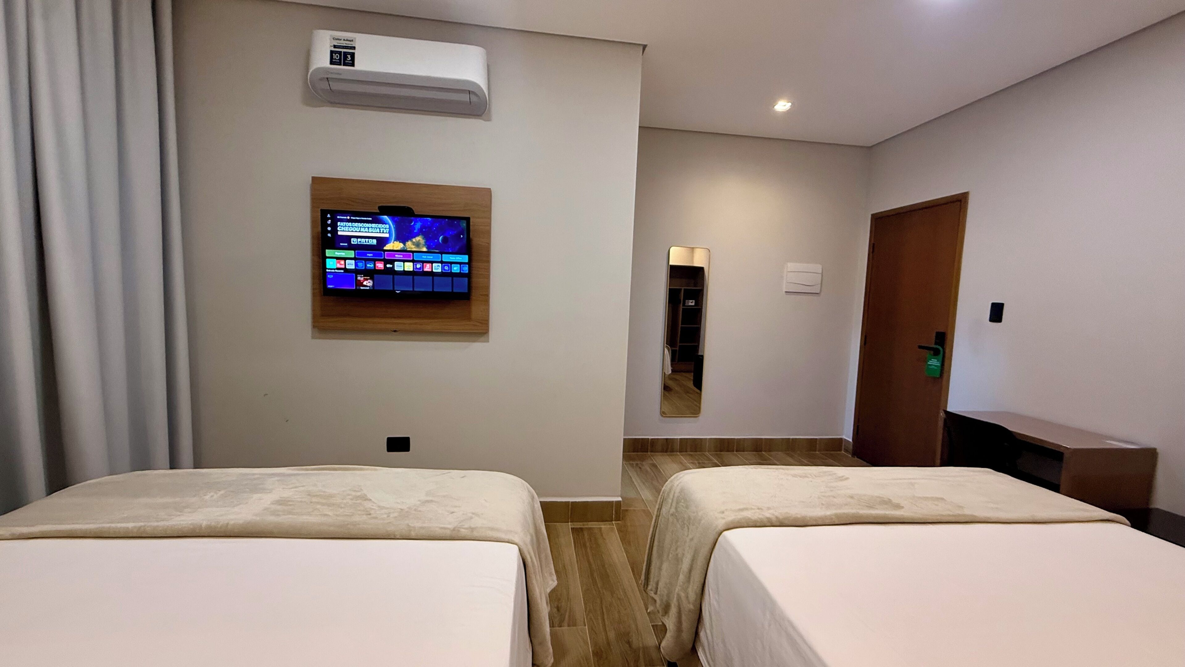 A hotel room with two beds, a TV mounted on the wall, an air conditioning unit, a mirror, and a door.