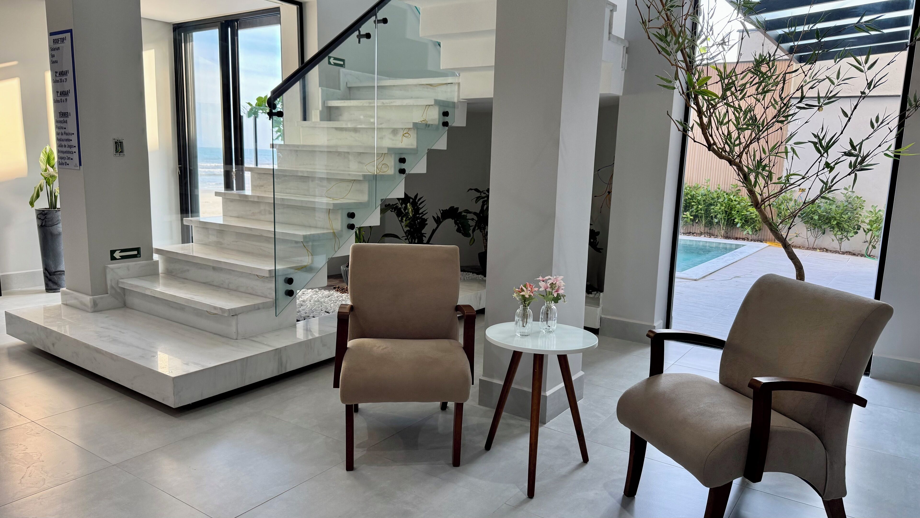 A modern interior with a glass staircase, two beige armchairs, a small round table with vases, and a view of the outdoors.