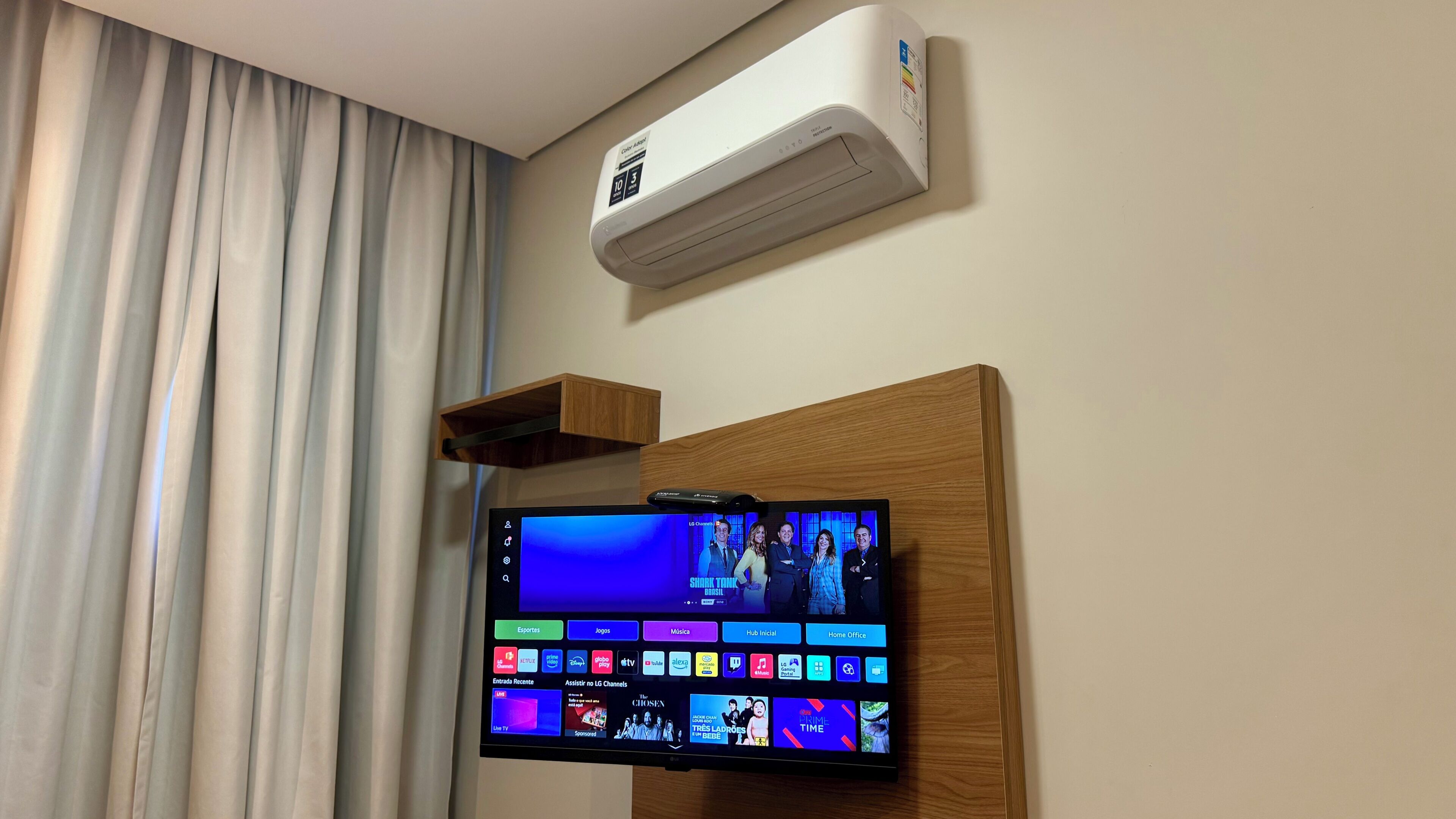 A hotel room with a mounted TV displaying various channels, a ceiling air conditioner, and a wooden shelf.