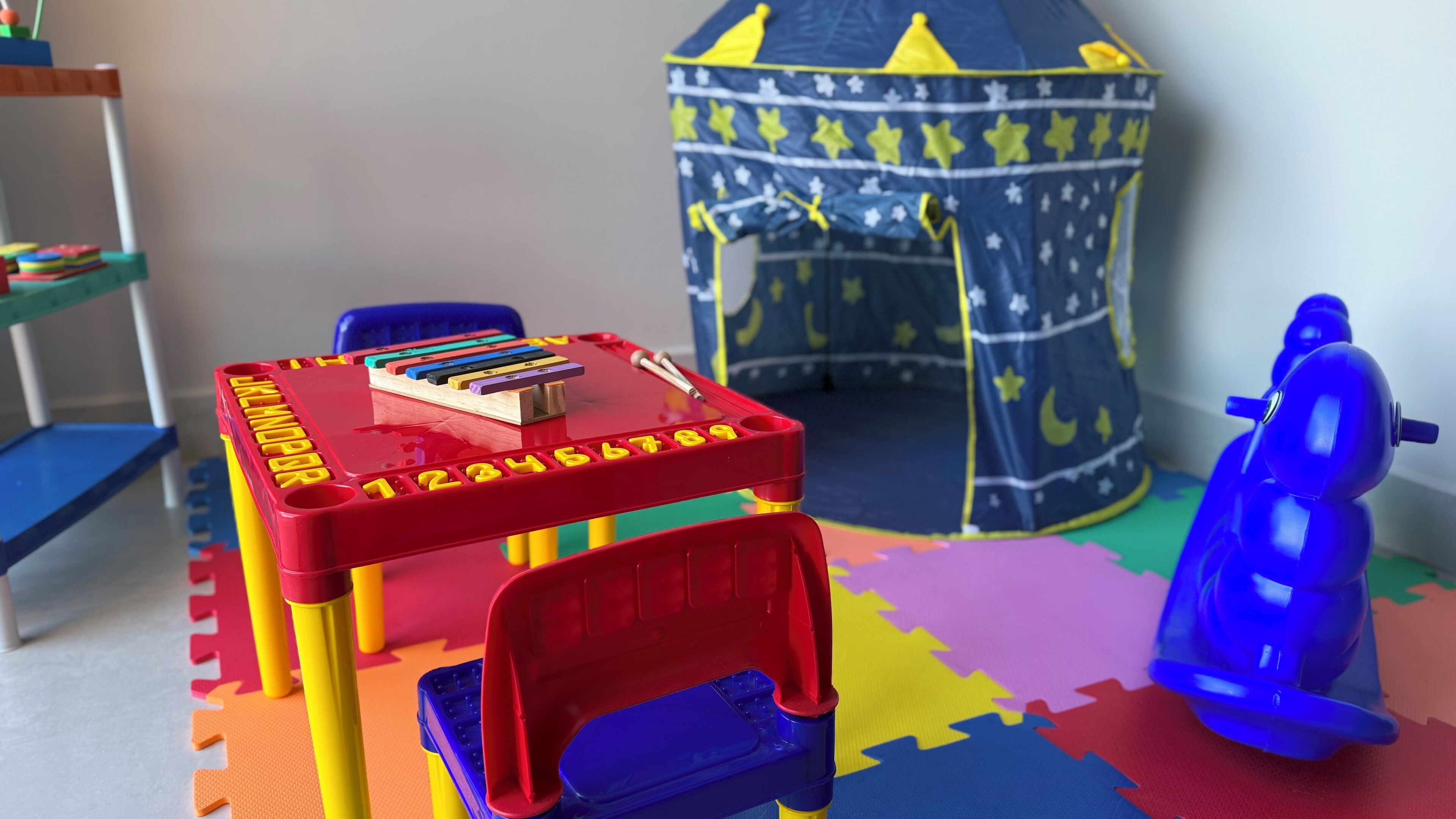 Uma área de recreação com uma mesa de brinquedo vermelha e azul, um carrinho de brinquedo azul e uma tenda de jogos com padrão de estrelas.