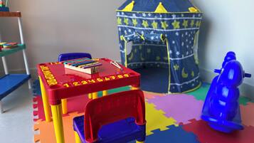 Uma área de recreação com uma mesa de brinquedo vermelha e azul, um carrinho de brinquedo azul e uma tenda de jogos com padrão de estrelas.