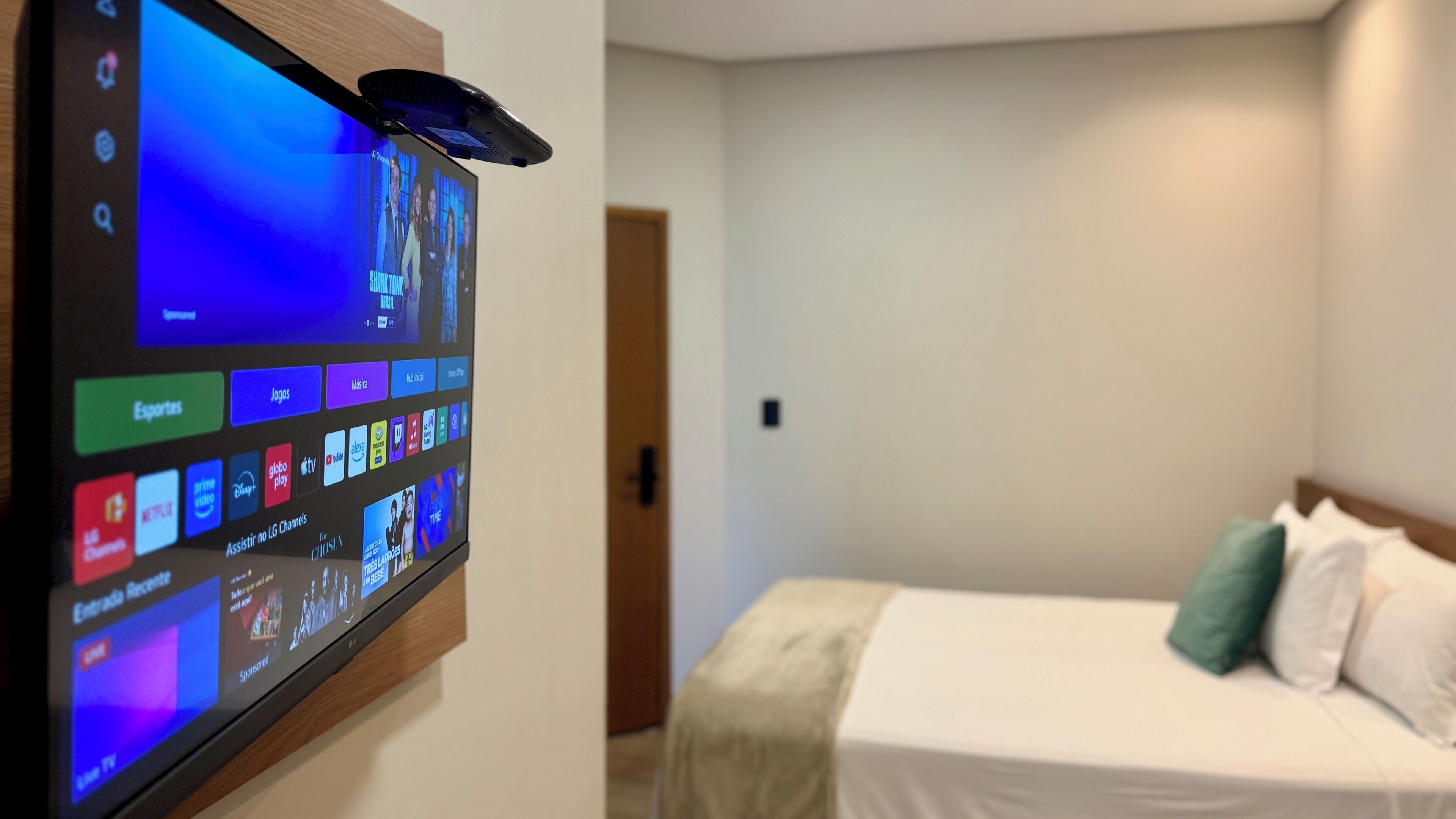 A hotel room with a mounted TV displaying a variety of channels and apps, a bed with pillows, and a wooden door.