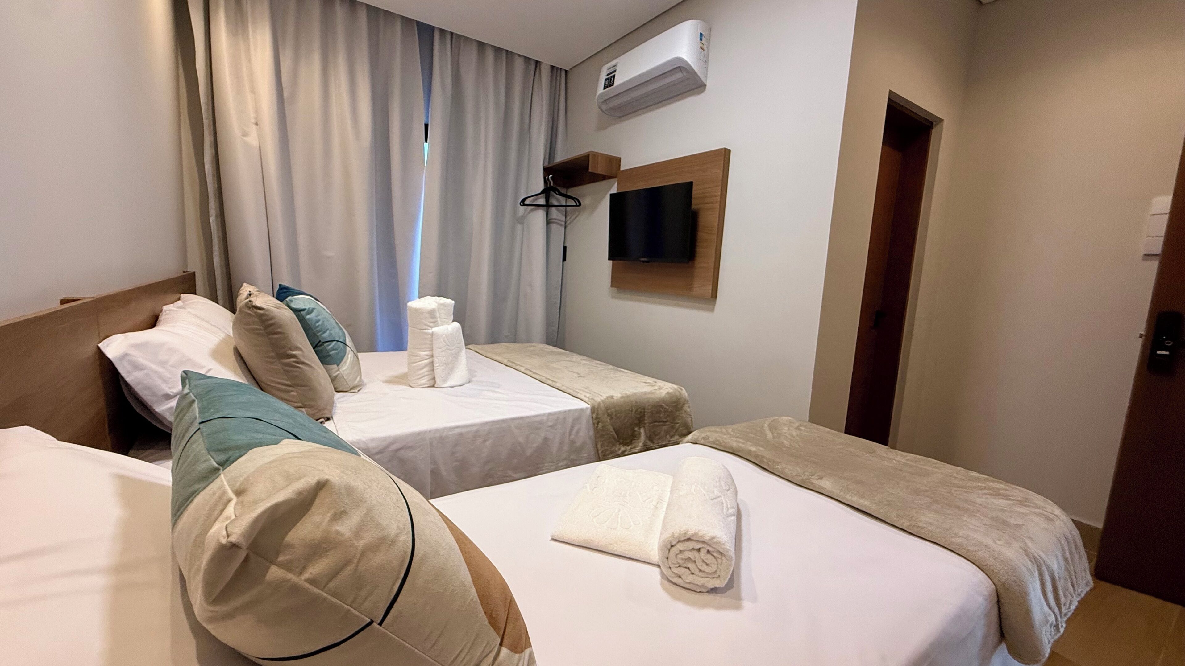 Quarto de hotel com duas camas de solteiro, televisão presa na parede, aparelho de ar condicionado e uma porta à direita.
