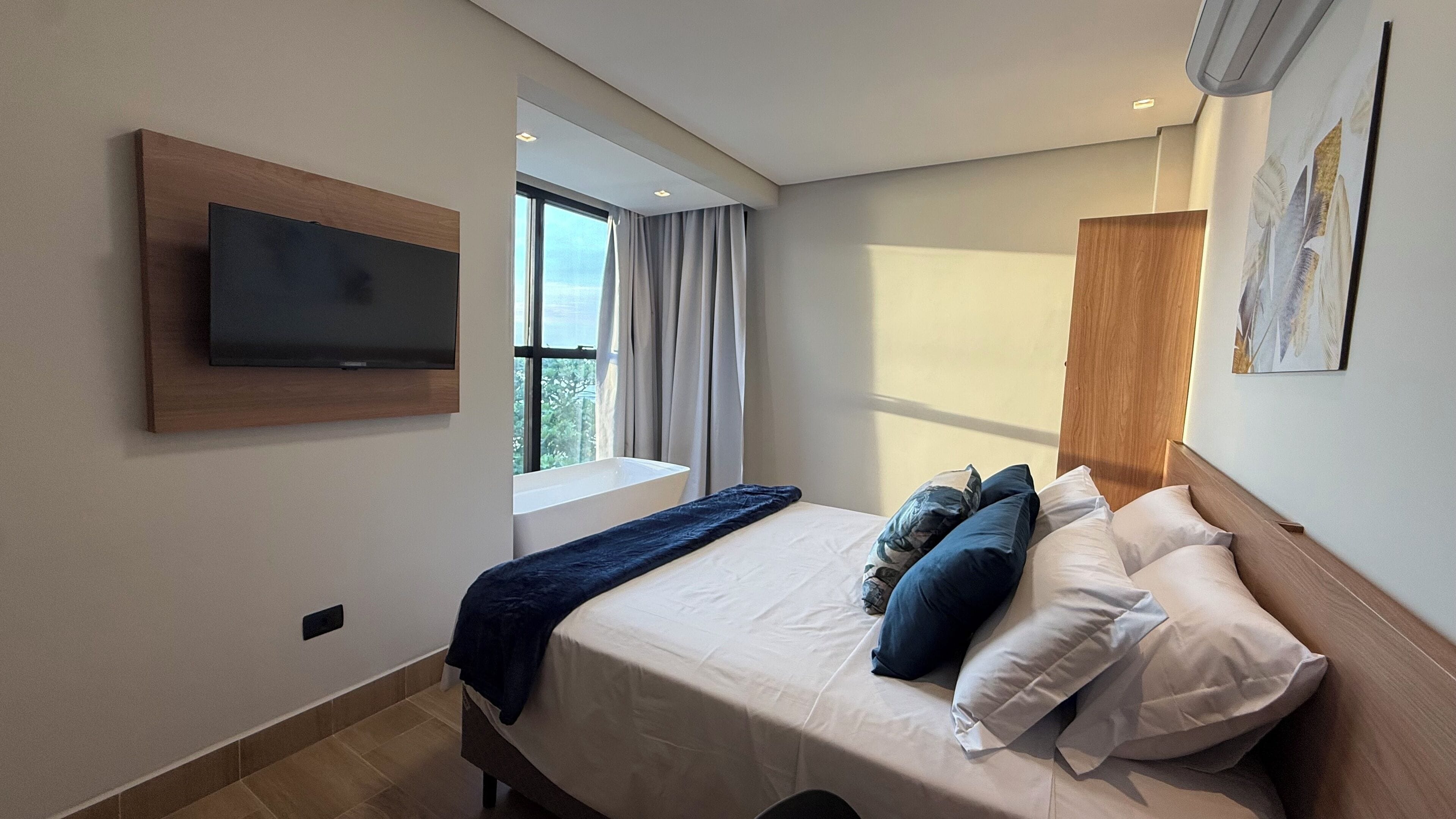 Una habitación de hotel moderna con una cama, un televisor montado, una ventana con cortinas y un cuadro en la pared.