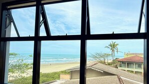 Vista pela janela mostrando uma praia e o oceano ao fundo, uma palmeira e um edifício com telhado vermelho.