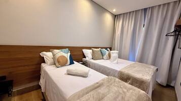 Quarto de hotel com duas camas, cada uma com roupa de cama branca e almofadas decorativas. Possui cabeceira de madeira, uma mesinha e uma cortina na janela.