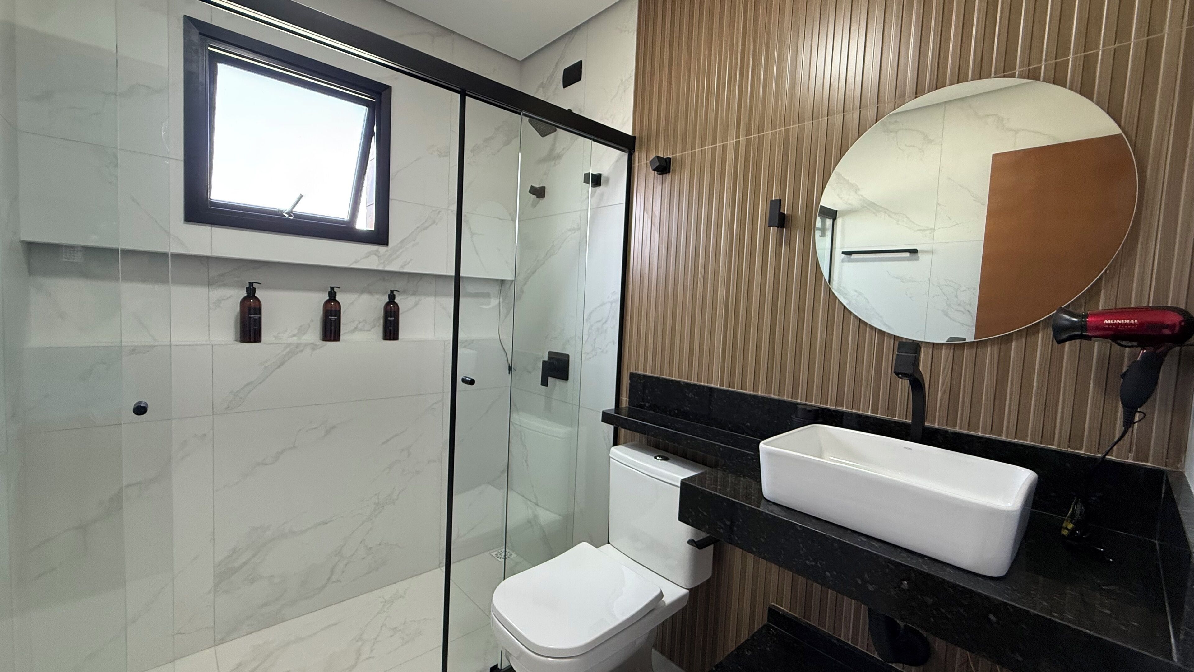 A modern bathroom with a glass shower enclosure, a white toilet, a black vanity with a white sink, and a round mirror.