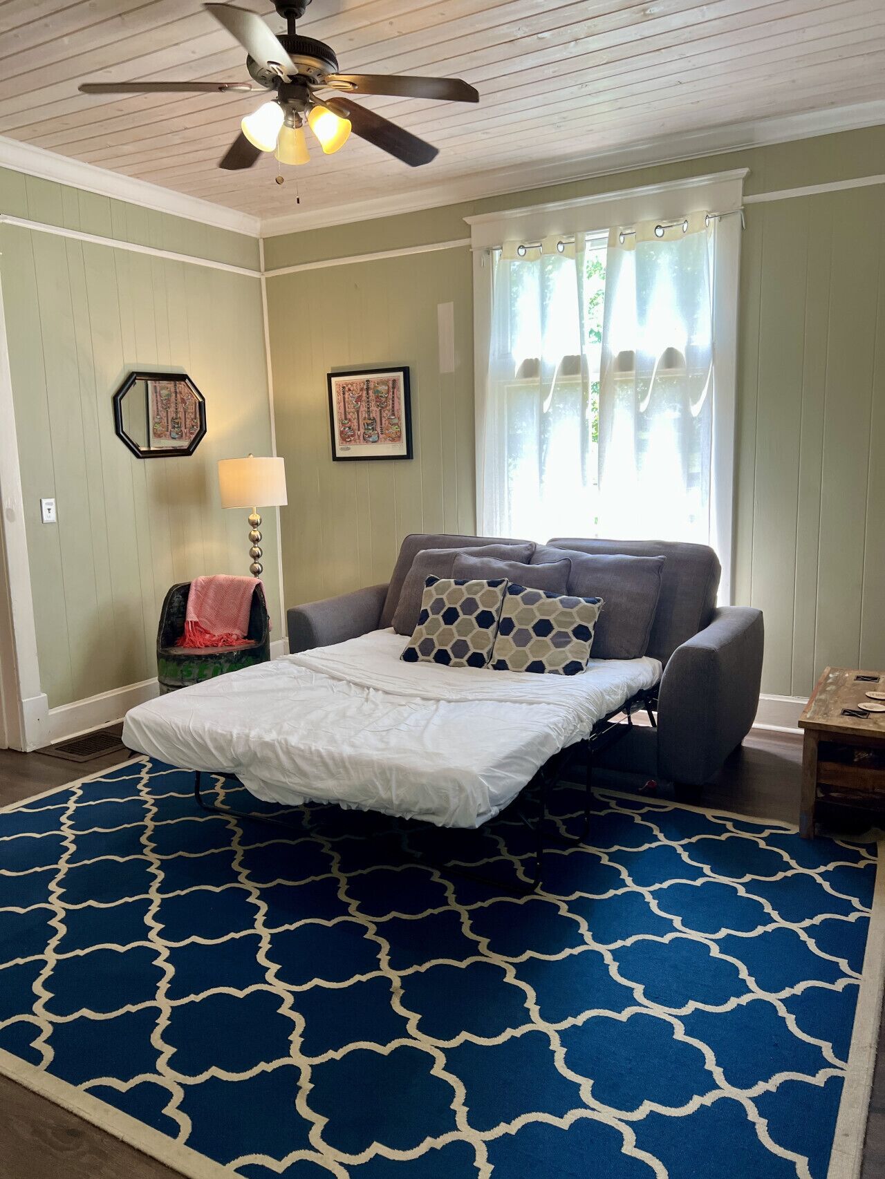 A bedroom with a bed, a ceiling fan, a window with curtains, and a blue patterned rug.