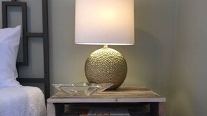 Une table de chevet avec une lampe au design original, une pile de livres et un lit blanc doté d’une tête de lit à motifs.