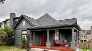 Une maison à deux étages avec une porte d’entrée rouge, un porche couvert avec des chaises et une cheminée en pierre.