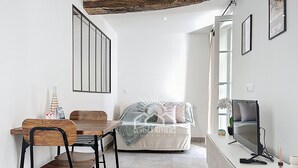 Una habitación pequeña y moderna con una mesa de comedor y sillas, una cama con colcha estampada, un televisor y una ventana con cortinas.
