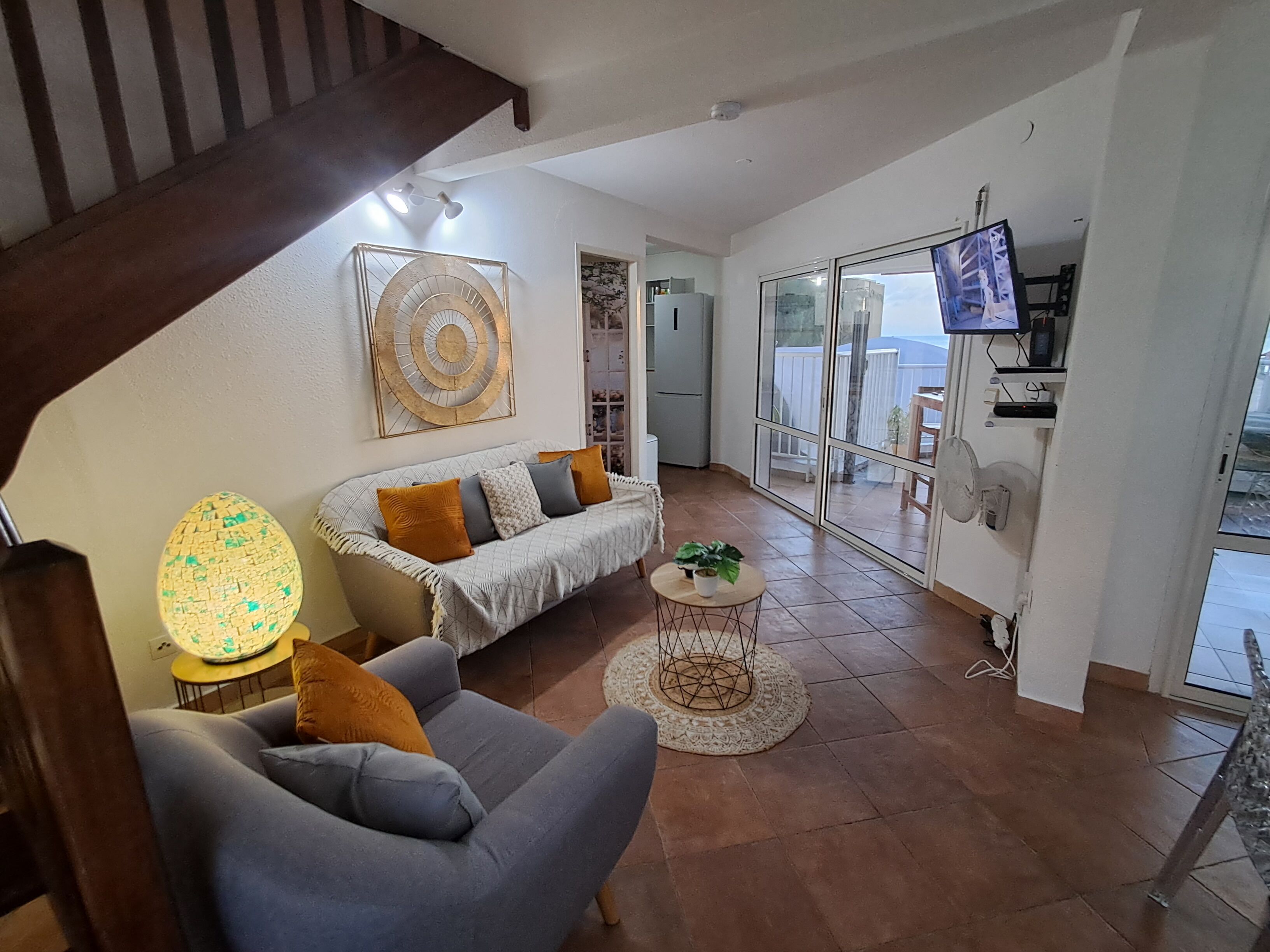 Un salon avec un canapé, un fauteuil et une table basse. Un téléviseur est fixé au mur, et on aperçoit un escalier au fond de la pièce.