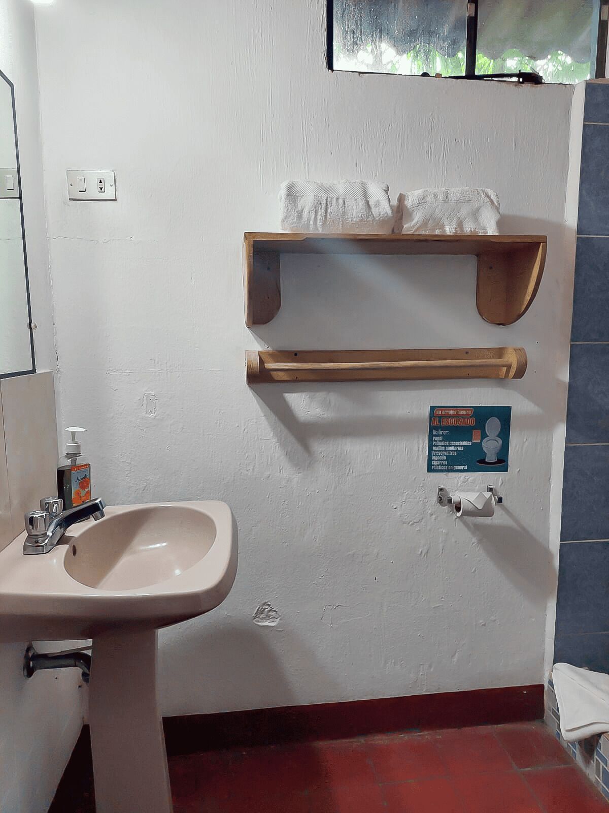 A bathroom with a sink, a soap dispenser, and a hand dryer. There are two wooden shelves above the sink, and a sign with instructions.
